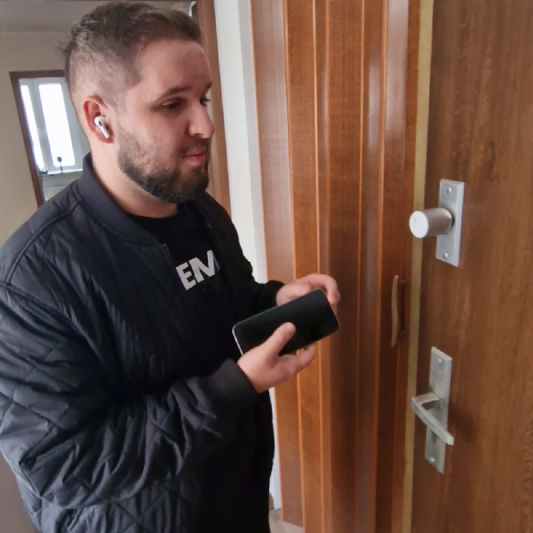 Een man staat bij een deur en gaat in interactie met een Tedee PRO slim slot dat op het oppervlak van de deur is gemonteerd. Hij houdt een smartphone in één hand, die hij gebruikt om het slot te bedienen. De man is Adrian Now, een online influencer met een visuele beperking die gespecialiseerd is in ondersteunende technologieën.